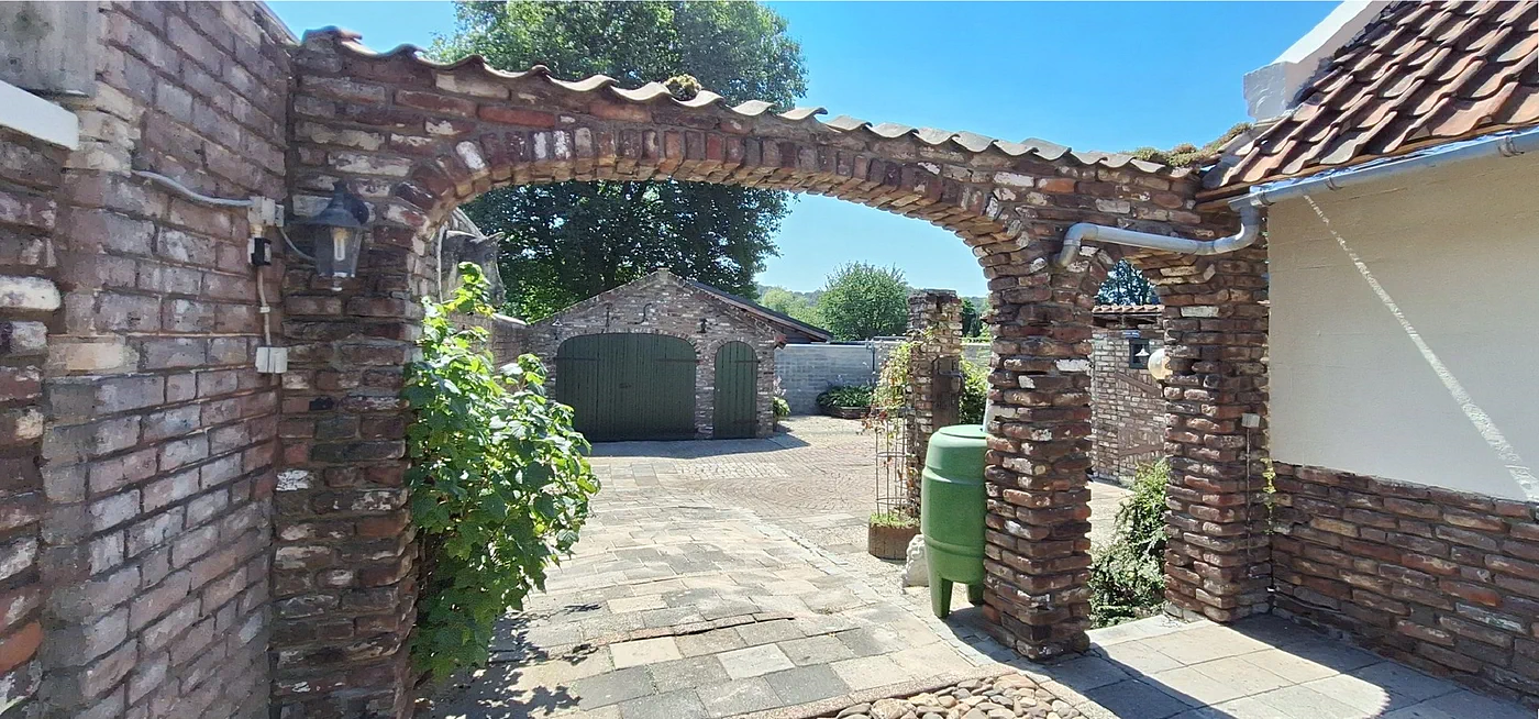 De gemetselde boog in de tuinmuur, met daarachter de tuin en de schuur met groene deuren