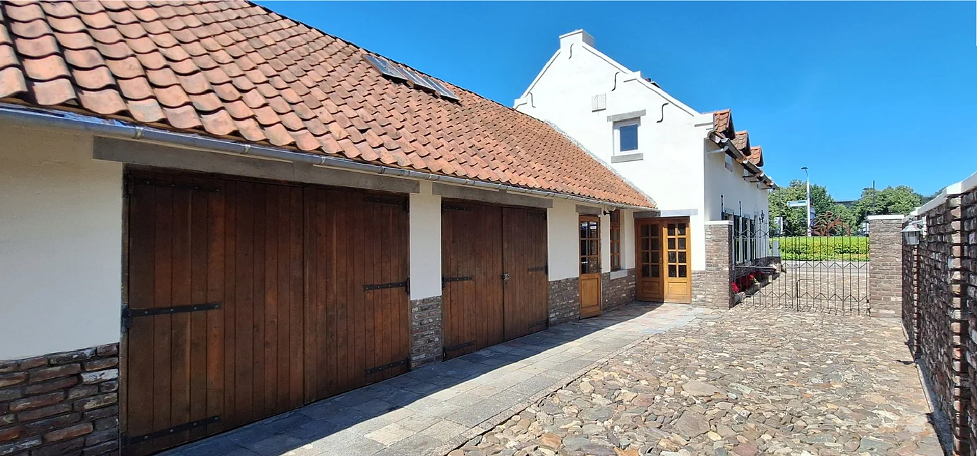 De bakstenen schuur met twee houten garagedeuren, met daarachter de witte puntgevel van het huis