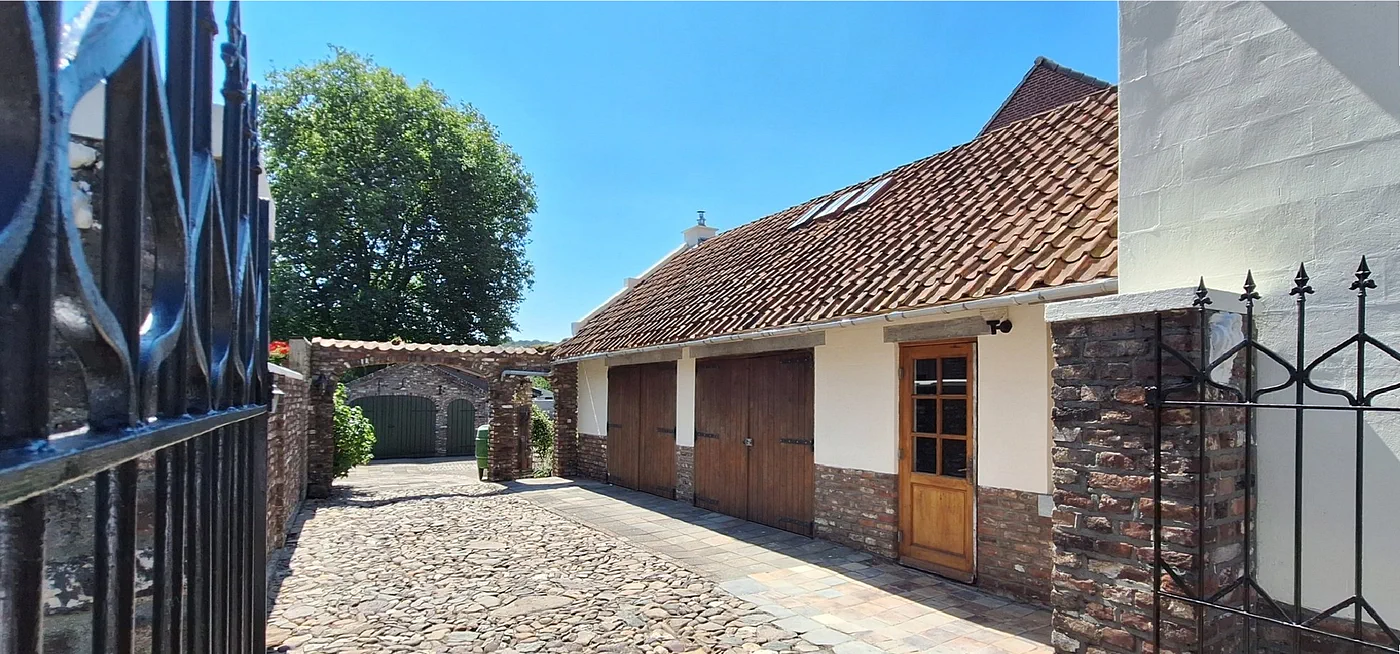 Door het smeedijzeren hek de oprit op: keien, de oude garagedeuren en de witte zijgevel van het huis
