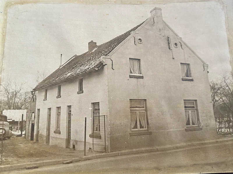 Oude foto van het huis: de mergelstenen gevel vlak langs de weg, met luiken, een hekje en een auto van toen