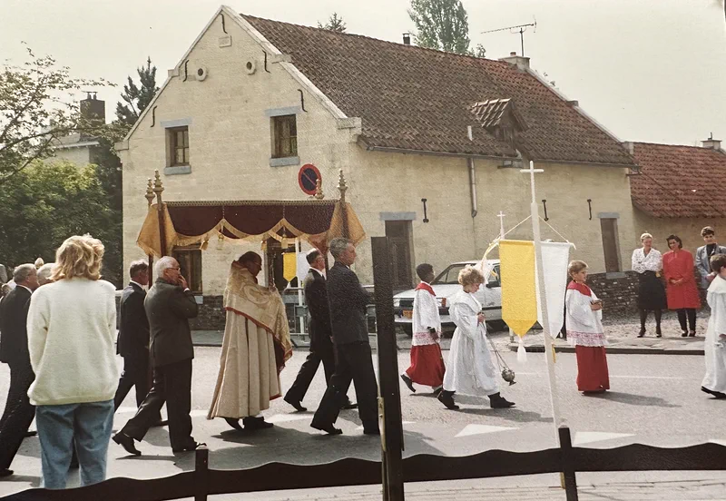 Een processie met misdienaars, vaandels en een priester onder een baldakijn trekt langs de gevel van het huis