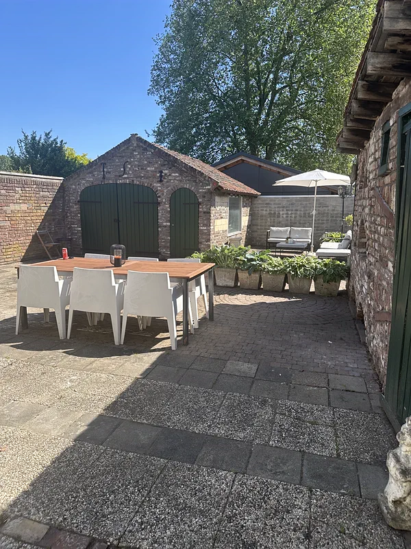 Het terras in de tuin: een houten tafel met witte stoelen op oude klinkers, met de schuur met groene deuren erachter